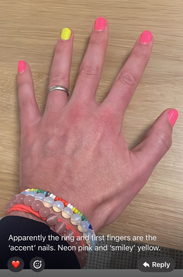 A hand resting on a desk with neon pink nails and one yellow. The participant wrote: Apparently the ring and first fingers are the accent nails. Neon pink and smiley yellow