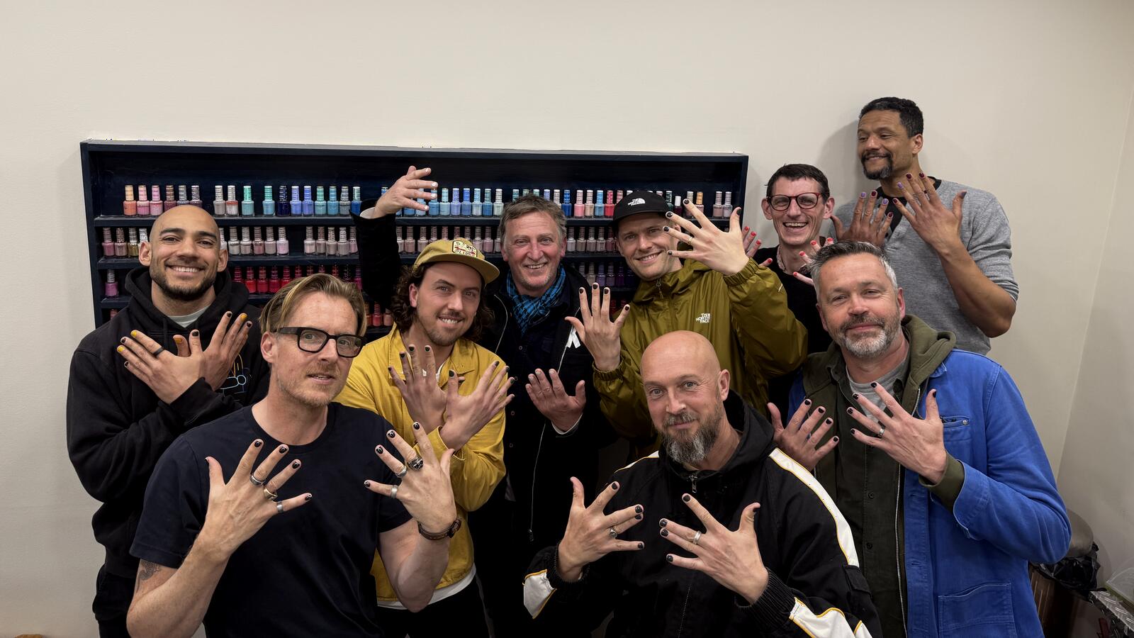 Nine smiling men in a nail salon holding up their freshly painted nails.