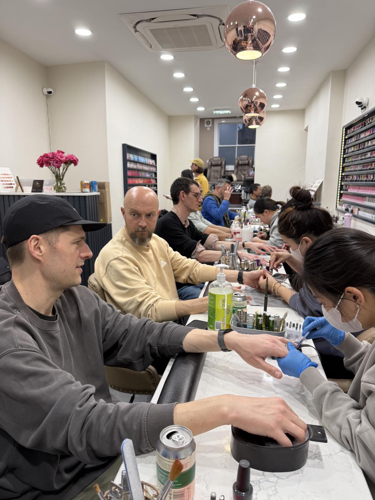 Men seated along a row of manicure tables in a salon, having their nails painted by technicians.