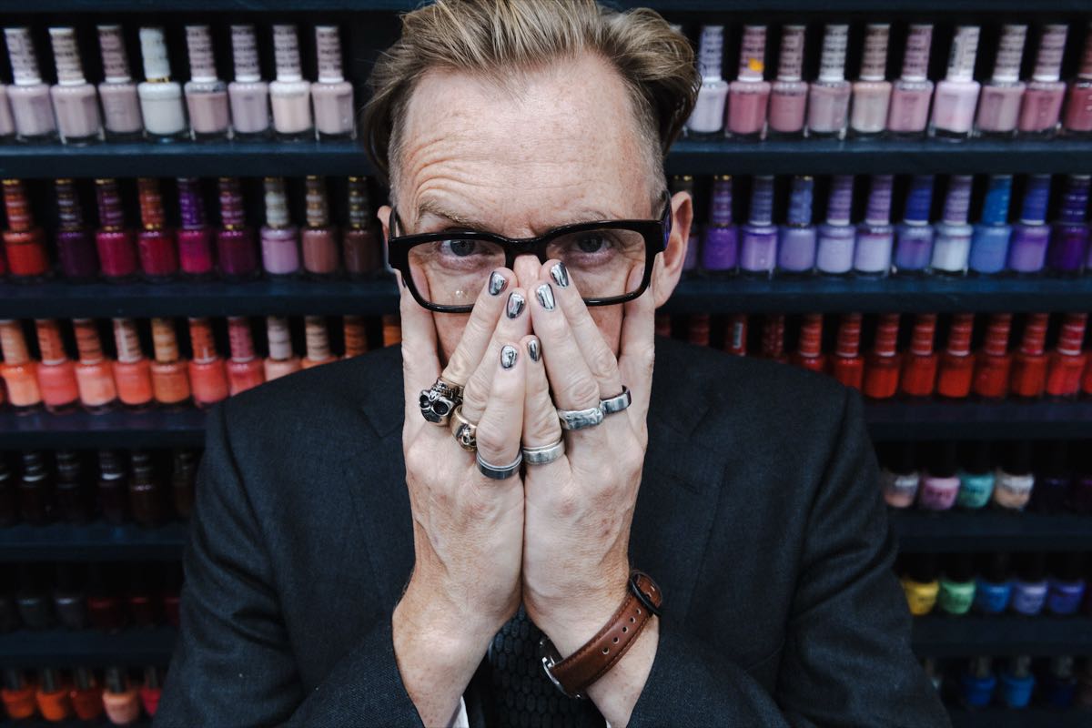 A man in a salon, hands raised, nails freshly painted, in front of a wall of colour.