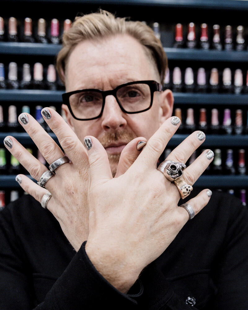 A man in glasses, hands crossed showing chrome painted nails and silver rings, in front of a wall of polish