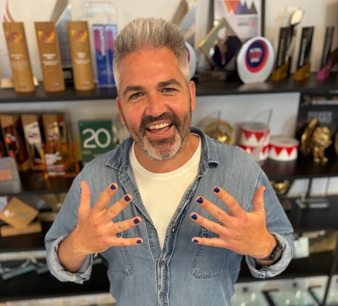 Dave Lowe in front of a shelf of industry awards, holding up his painted nails